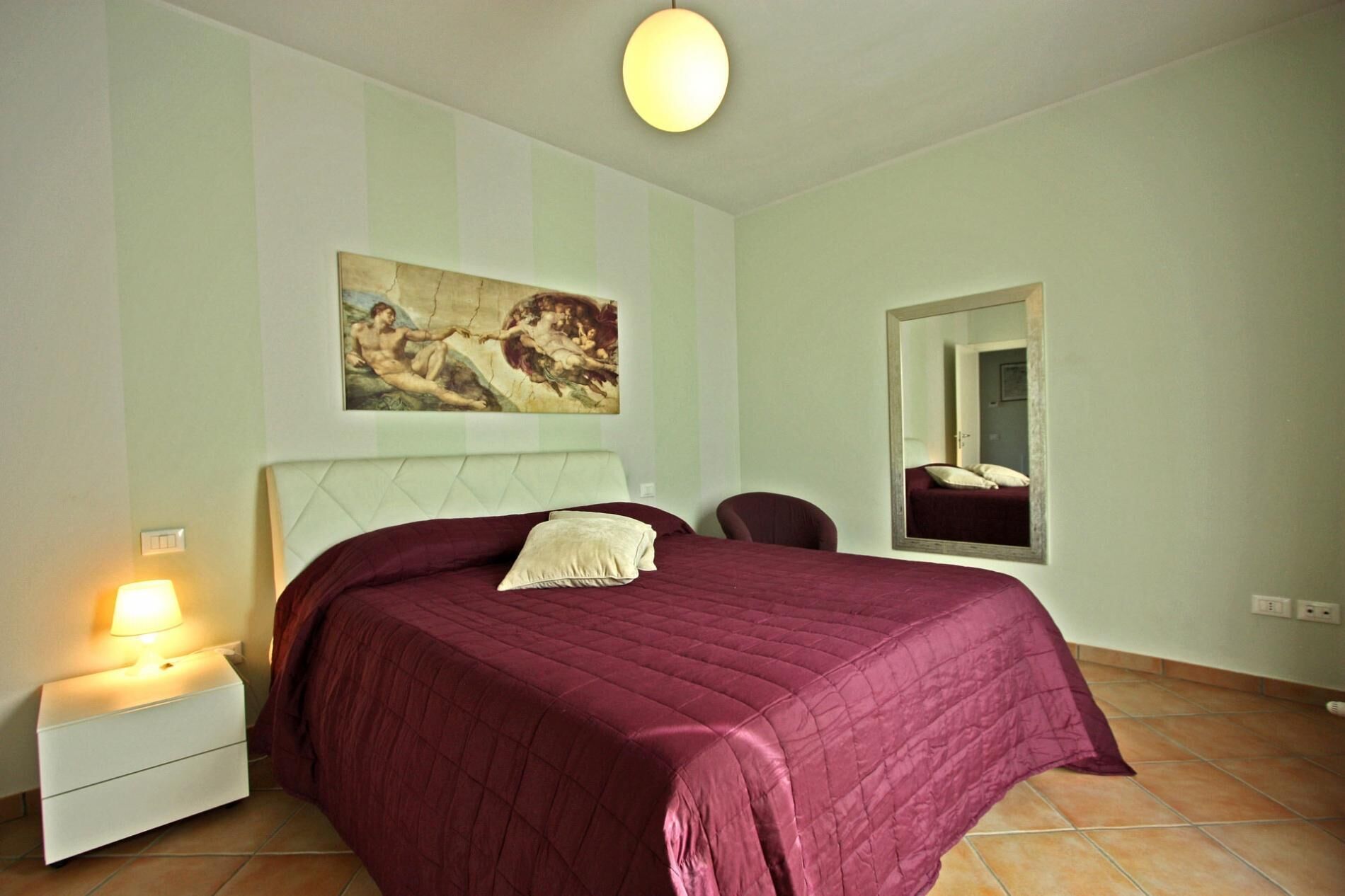 A bedroom with a large bed, a nightstand with a lamp, a chair, a painting on the wall, and a mirror.