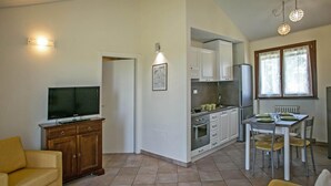 Un espace de vie compact comprenant un téléviseur, un meuble en bois, une table à manger, des chaises, un coin cuisine et une fenêtre avec des rideaux.
