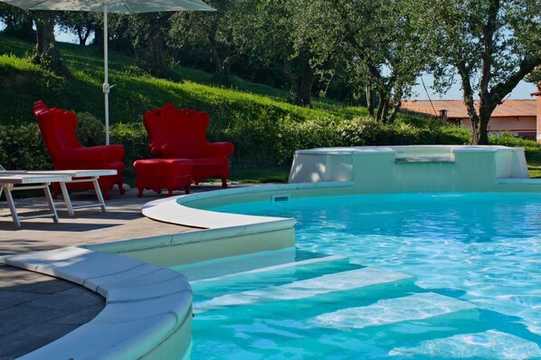 Una piscina con bordo curvo, circondata da un'area verde e alberi. Sono presenti sedie rosse e un tavolo vicino alla piscina.