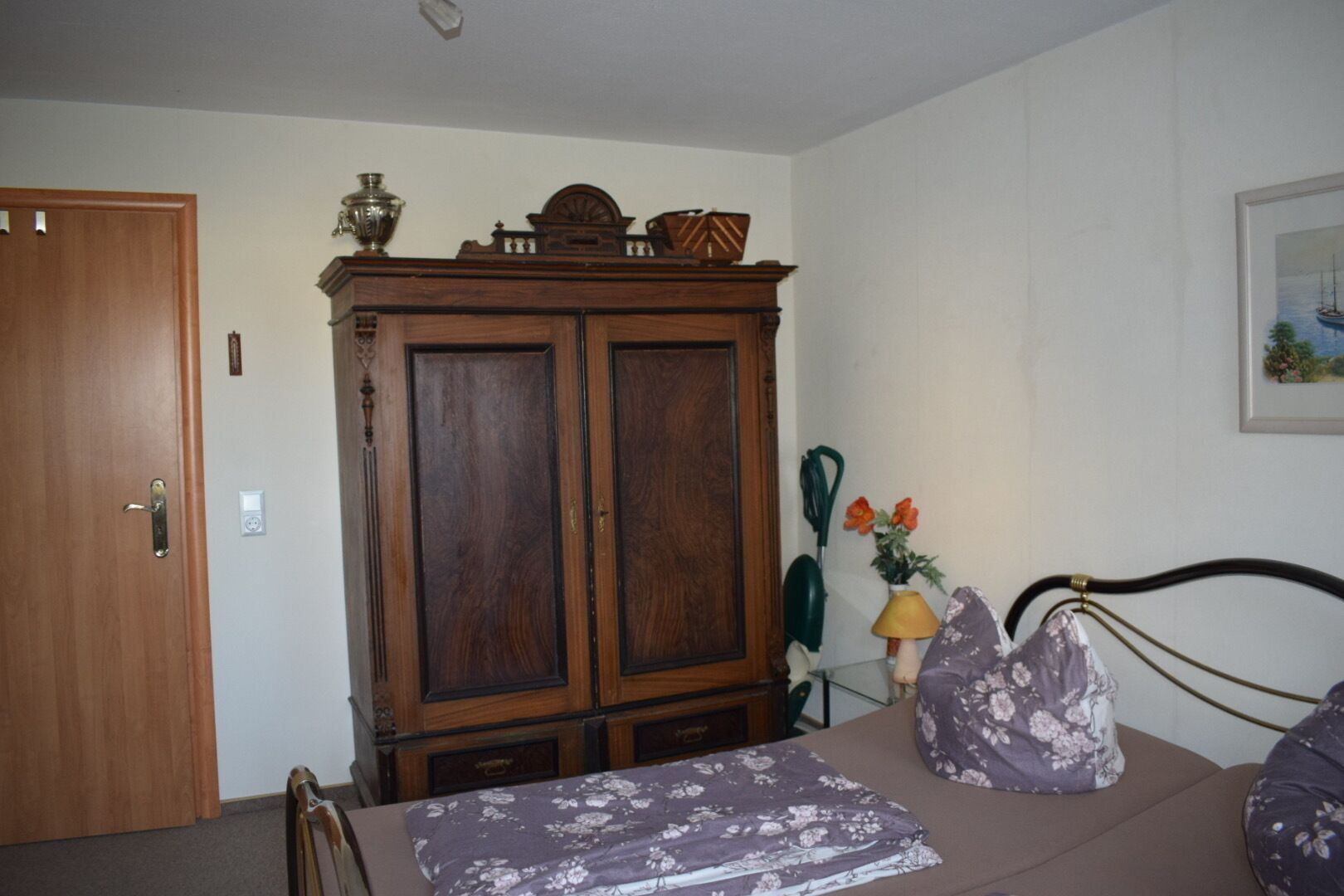Een slaapkamer met een houten garderobe, een bed met een bloemenbedekking, een nachtkastje met een lamp en een lijst met een foto aan de muur.