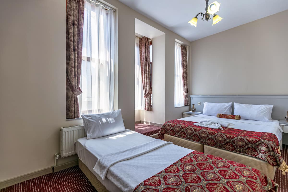 A hotel room with two beds, a radiator, a window with curtains, a nightstand, and a chandelier.