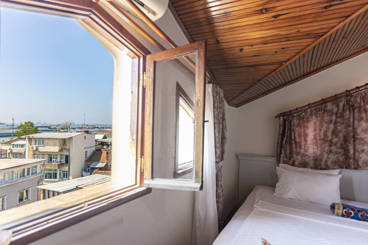 A bedroom with a large window, a wooden ceiling, and a view of buildings.