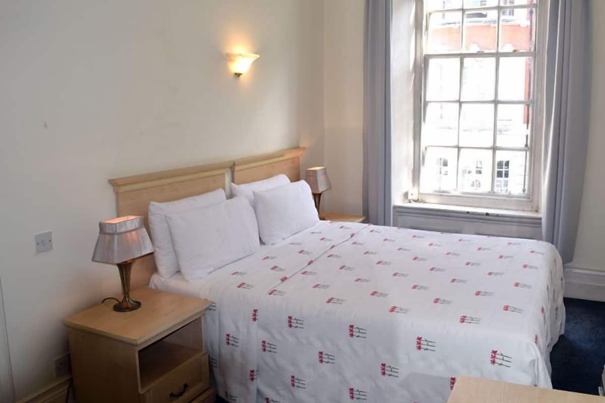A hotel room with a bed, bedside lamps, a nightstand, a window with a view of buildings, and a wall-mounted light.