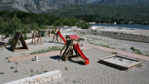 Ein Spielplatz mit Schaukeln und Rutschen, einer kleinen Schutzhütte und einem Sandspielbereich, vor einer Bergkulisse und einem Gewässer.