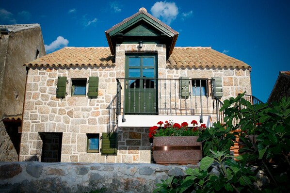 A stone building with a tiled roof, a balcony with a metal railing, and a large planter with red flowers.