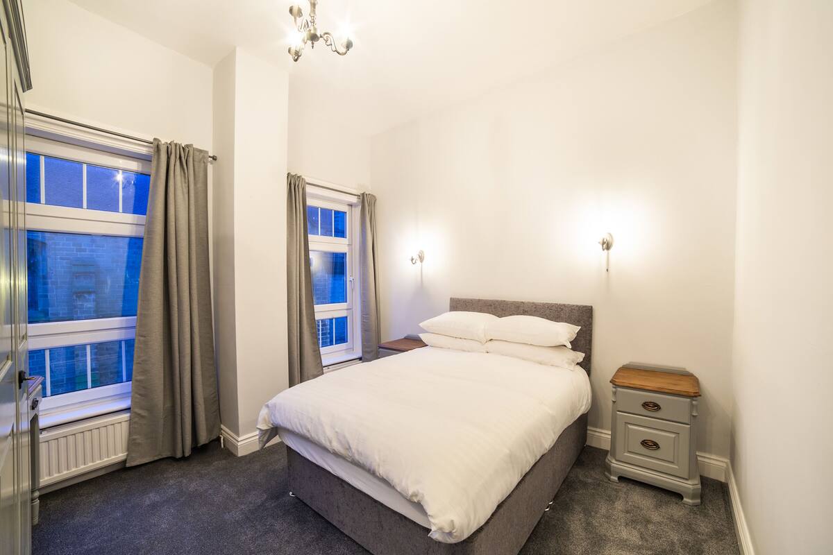 A bedroom with a bed, a nightstand, a dresser, and two windows with curtains.