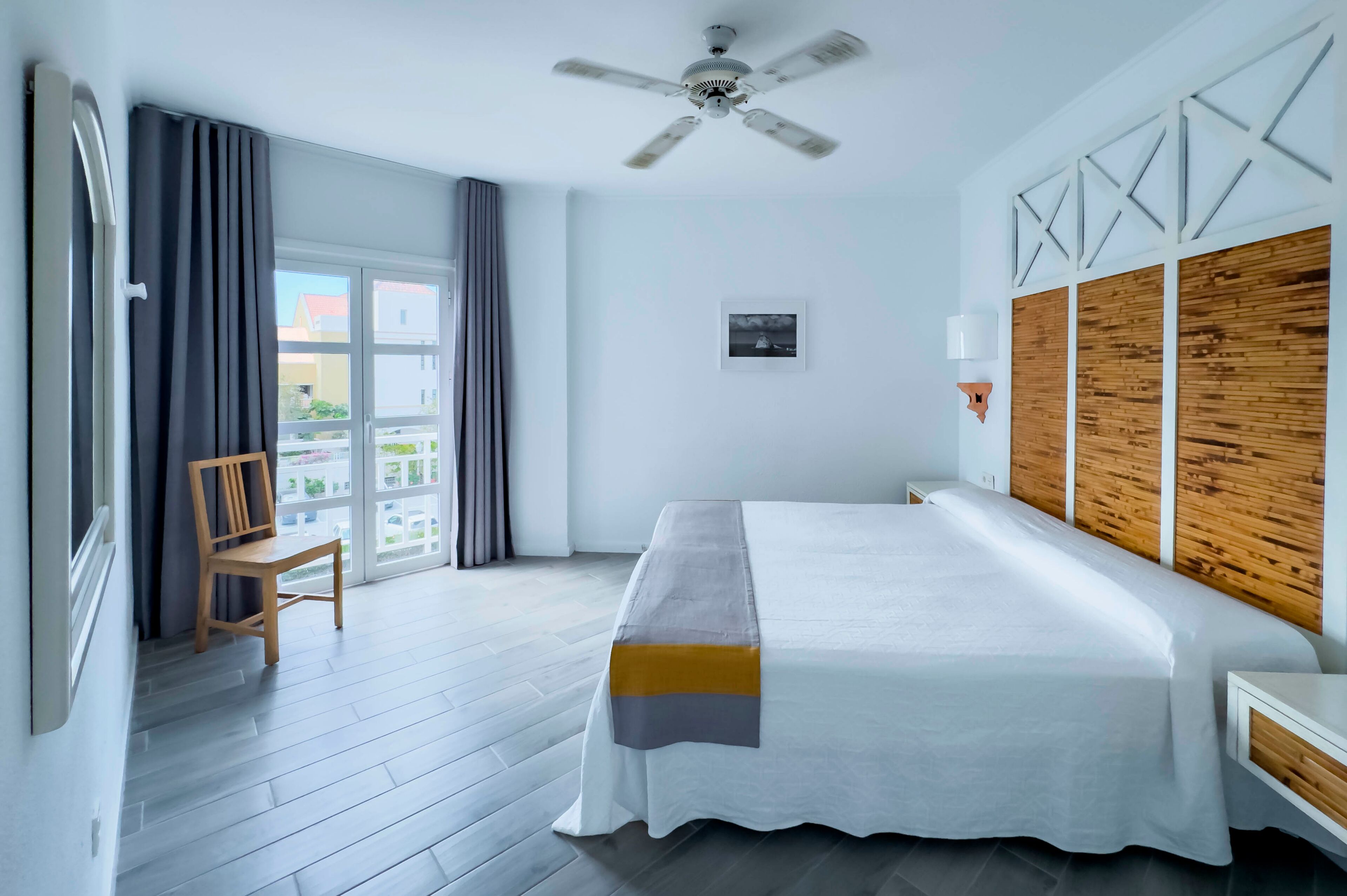 Ein Hotelzimmer mit einem großen Bett, einem Deckenventilator, einem Holz-Kopfteil, einem Fenster mit Vorhängen, einem Sessel und Blick auf Gebäude.