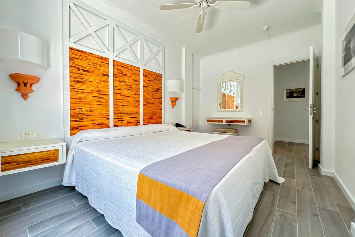 A bedroom with a large bed, wooden panel wall, white decor, and a ceiling fan.