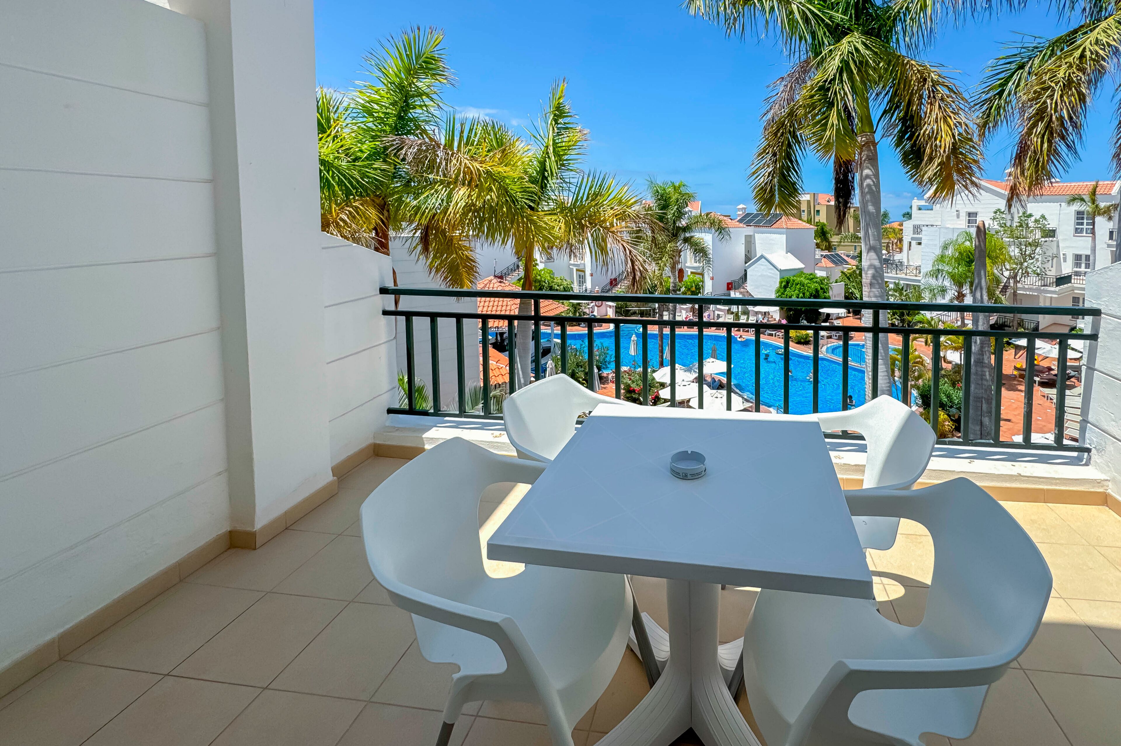 Un balcón con una mesa y sillas blancas, con vista a una piscina y palmeras.