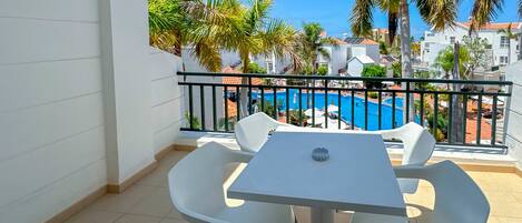 Un balcón con una mesa y sillas blancas, con vista a una piscina y palmeras.