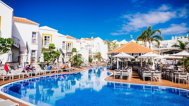 A swimming pool with lounge chairs and umbrellas, surrounded by white buildings with orange roofs. - Apartamentos Parque del Sol (Adeje)