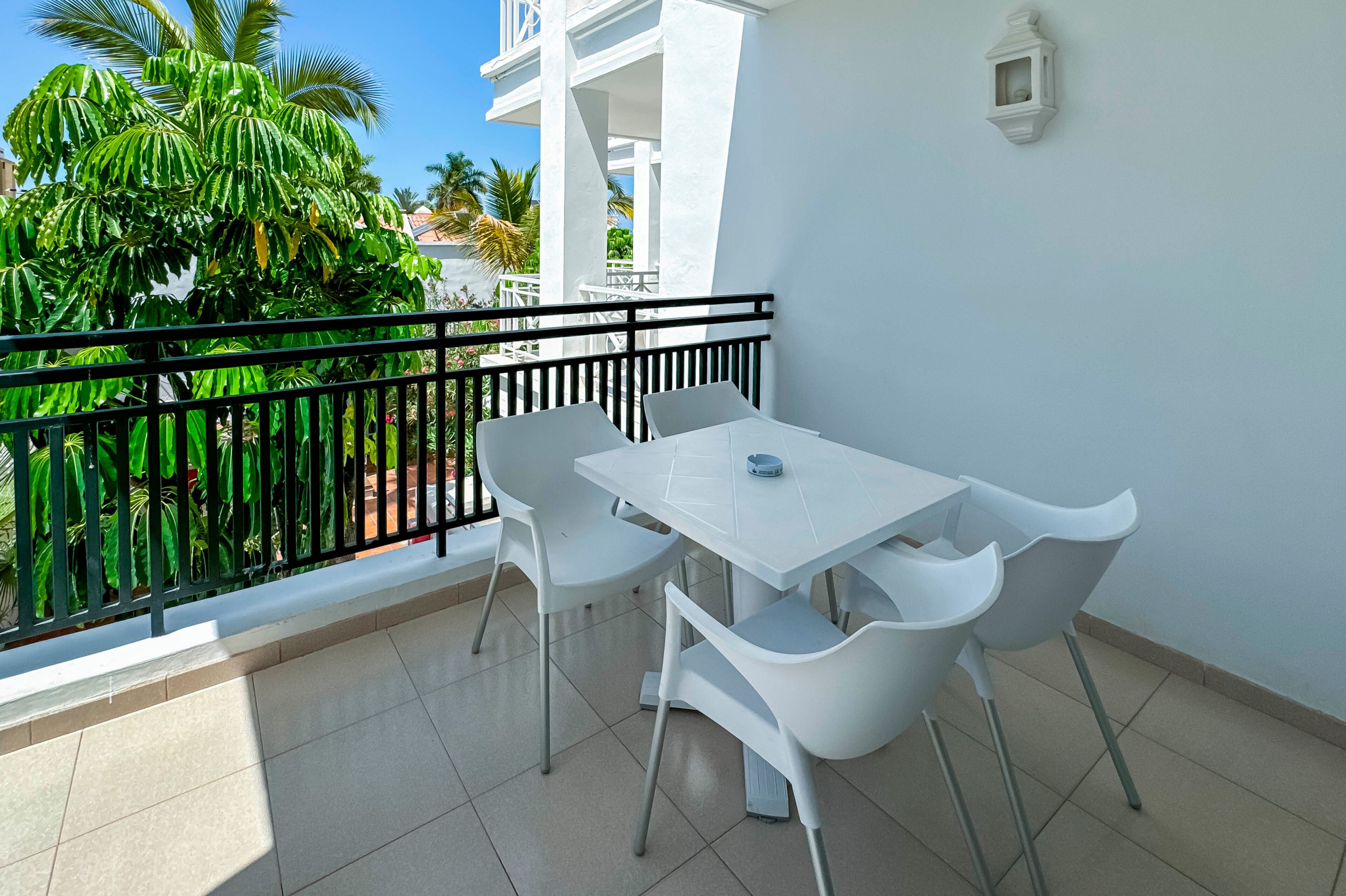 A balcony with a white table and chairs, a black railing, and a view of greenery and a building in the background.
