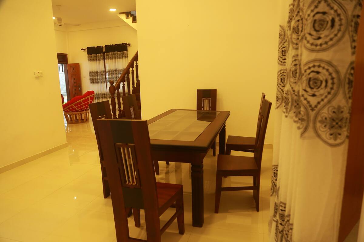 Un comedor con una mesa y sillas de madera, una escalera al fondo y una cortina estampada a la derecha.