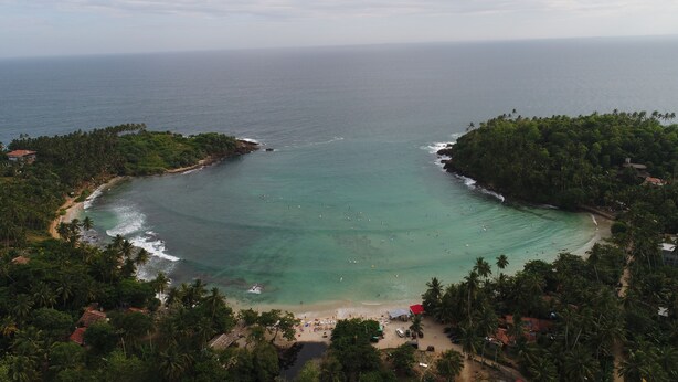 An aerial view of a tropical beach with clear turquoise waters, surrounded by dense palm trees and a few buildings.
