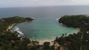 An aerial view of a tropical beach with clear turquoise waters, surrounded by dense palm trees and a few buildings.