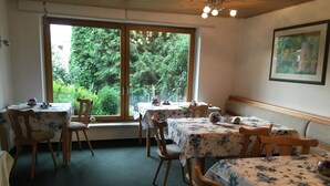 A dining area with wooden chairs and tables, large windows with a view of greenery, and a ceiling light fixture. - Pension Anna Gastronomie (Loebau)