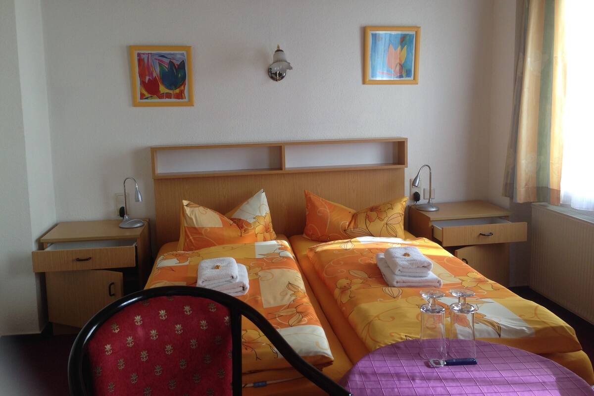A hotel room with two beds, a wooden headboard, bedside tables with lamps, a chair, a small table with a glass, and artwork on the walls.