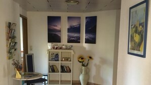 A hallway with a tiled floor, a white bookshelf, a round table with a chair, and a vase with sunflowers. - Pension Anna Gastronomie (Loebau)