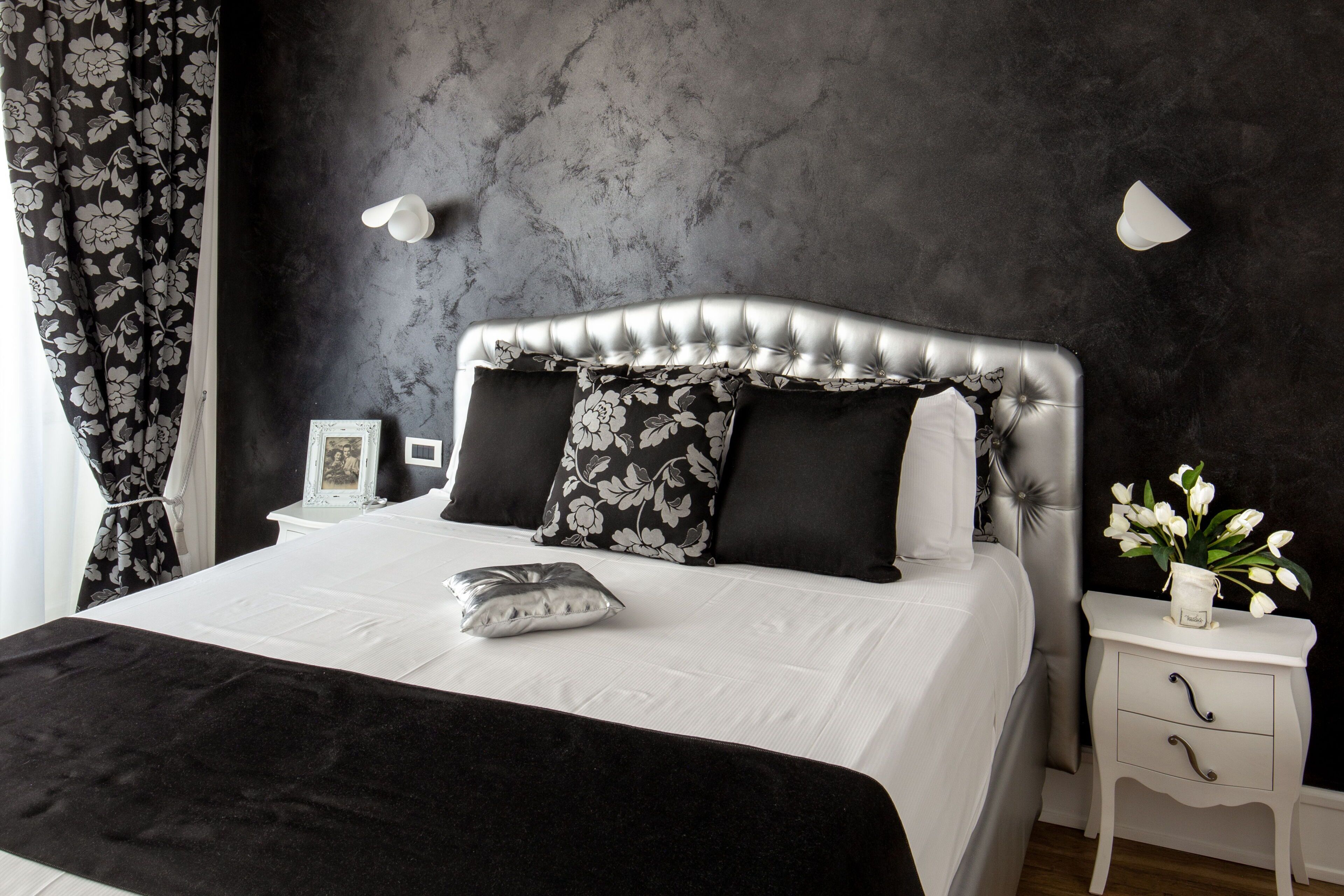 A bedroom with a tufted headboard, white bedding, black and white floral pillows, a bedside table with a vase of flowers, and a dark textured wall.