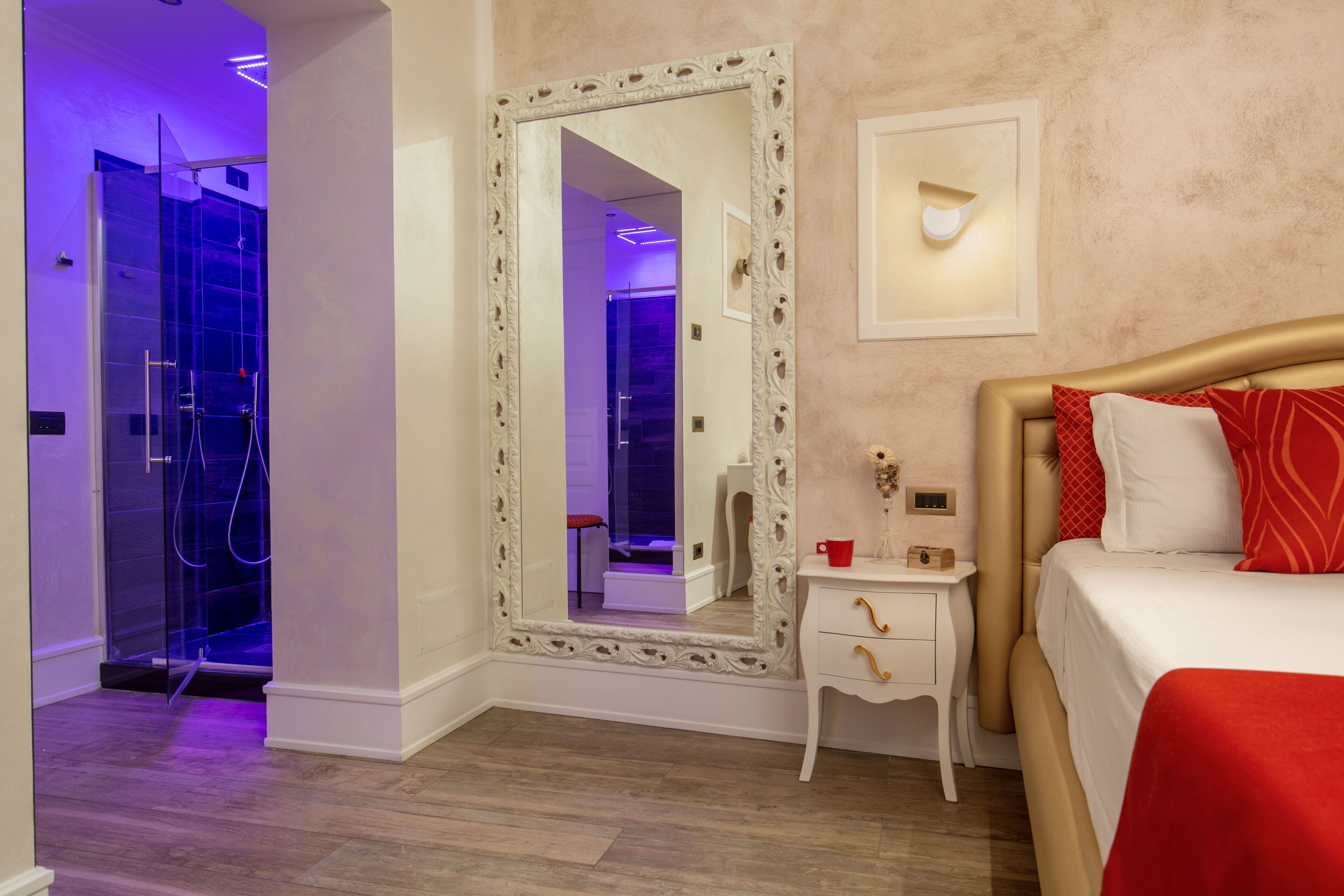 A hotel room with a large mirror, a bed with red bedding, a bedside table with a red cup, and a shower with blue lighting.