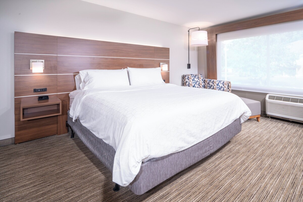 A modern hotel room with a large bed, a nightstand, a lamp, and a window with a view.