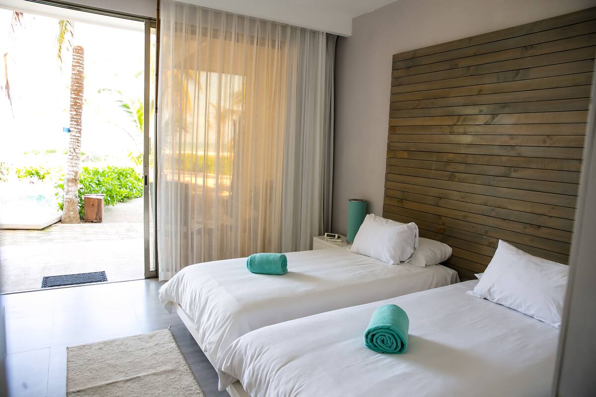 A hotel room with two beds, a wooden headboard, a large window with sheer curtains, and a view of the outdoors.