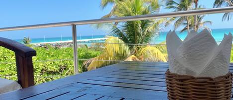 Una mesa con una cesta de mimbre que contiene servilletas, un tenedor y una cuchara, con vistas a una playa con palmeras y un cielo azul despejado.