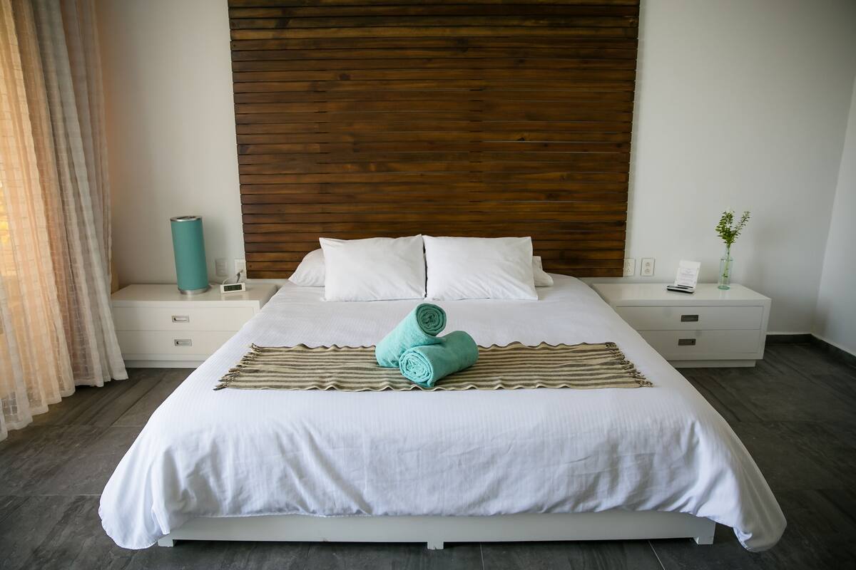 A neatly made bed with white linens and rolled towels, a wooden headboard, and a bedside table with a lamp and a small plant.