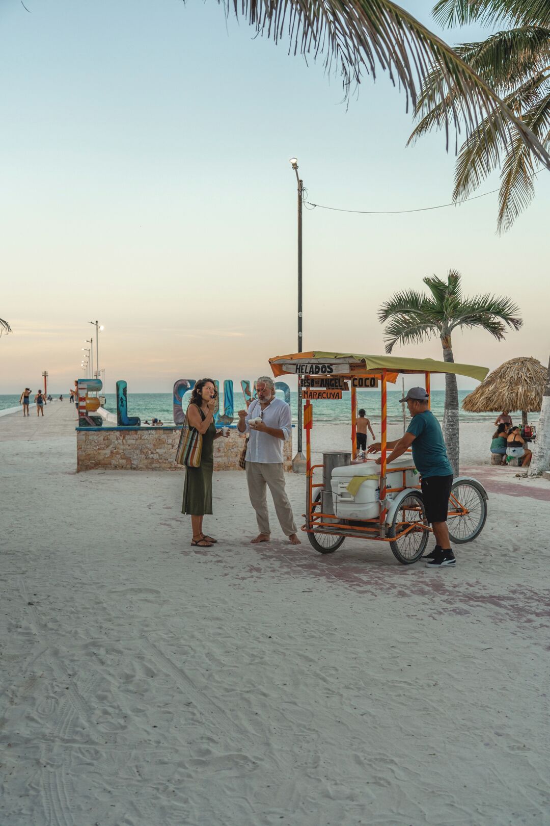 Cena de praia com um carrinho identificado como 'HELADOS BESOS DE ANGEL MARACUYÁ' e palmeiras.