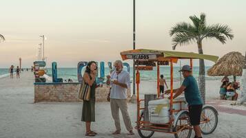 Cena de praia com um carrinho identificado como 'HELADOS BESOS DE ANGEL MARACUYÁ' e palmeiras.