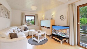 A living room with a white sofa, a TV on a stand, a small table, and a window with curtains. - Holiday house near Anne (sauna, forest, lake) (Kirchheim)