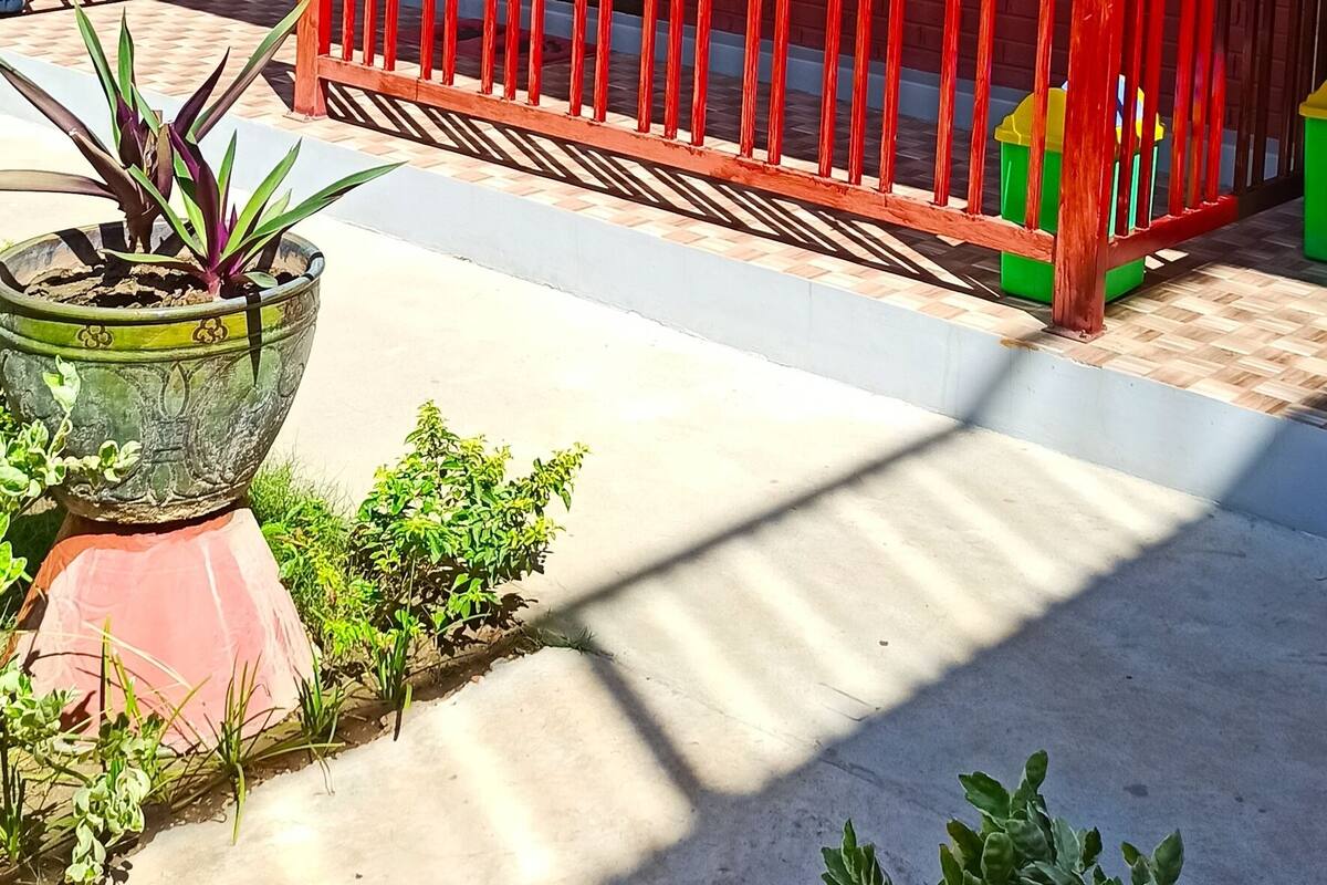 A red building with a wooden door, a balcony with red railings, and potted plants on the ground.