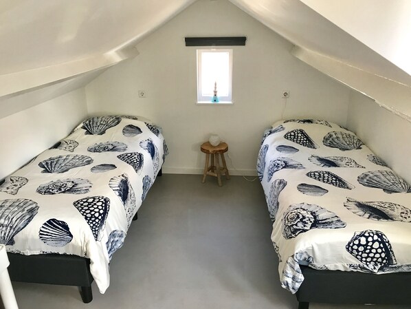 Une petite chambre aménagée sous les toits, avec deux lits simples, chacun recouvert d’une couvre-lit à motifs de coquillages. On y trouve un petit tabouret en bois et une fenêtre donnant sur le ciel.