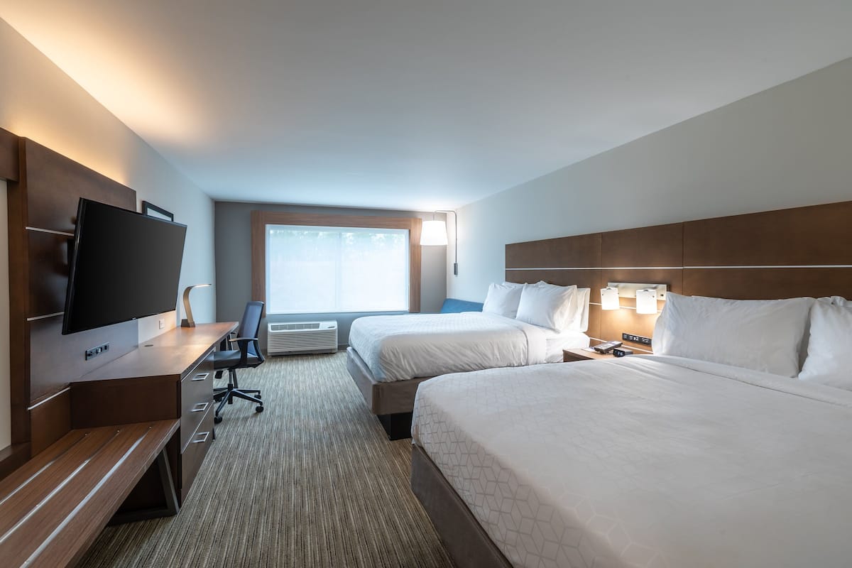 Quarto de hotel com duas camas, uma escrivaninha com televisão de tela plana, uma cadeira e uma janela grande com persianas.