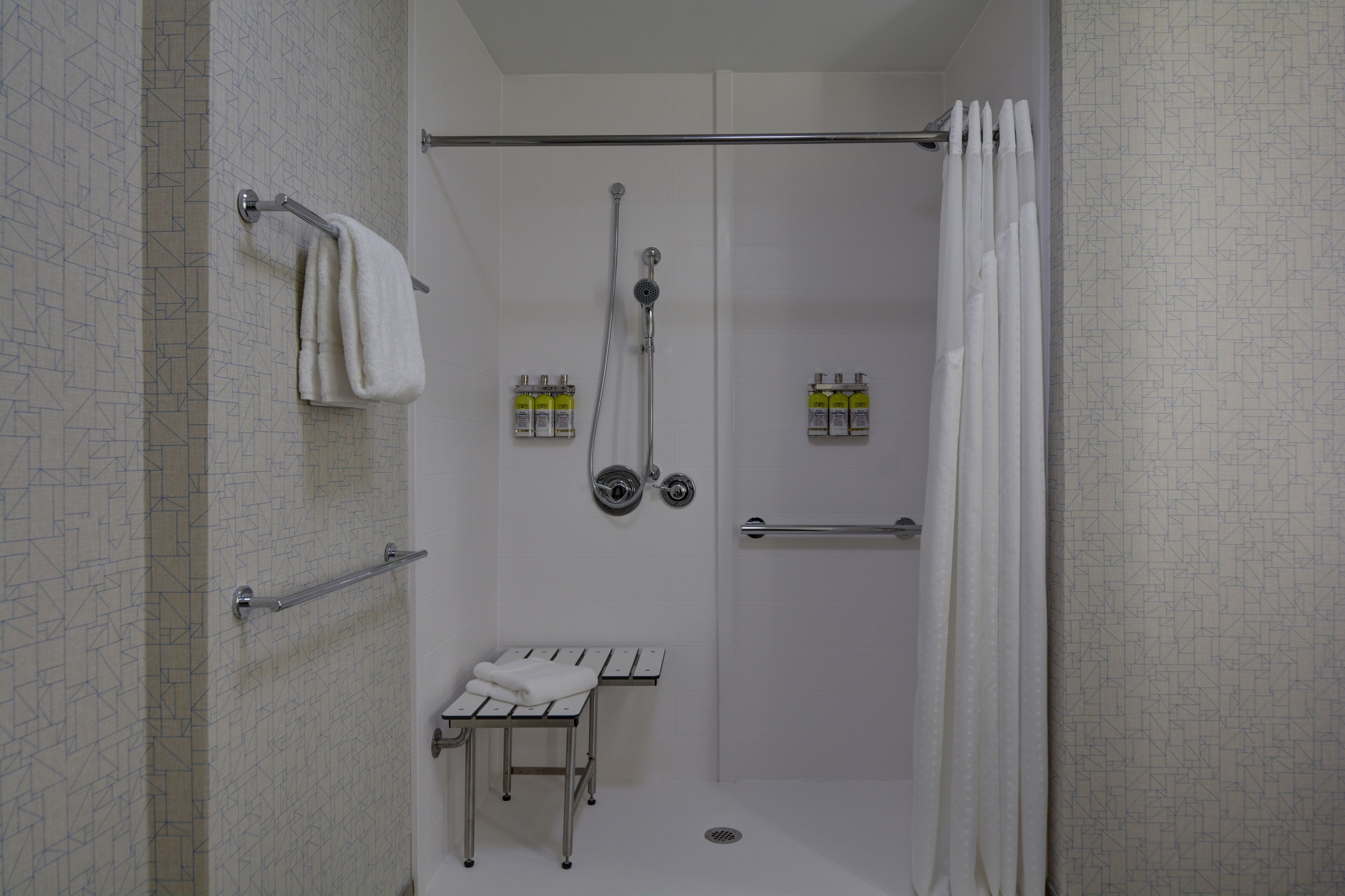 Een inloopdouche met een glazen deur, een handdoekhouder, een bankje en twee flessen shampoo.