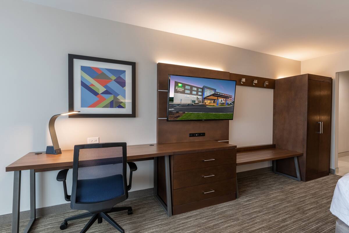 Quarto de hotel com uma escrivaninha de madeira, uma cadeira, uma televisão fixada na parede e um quadro emoldurado.