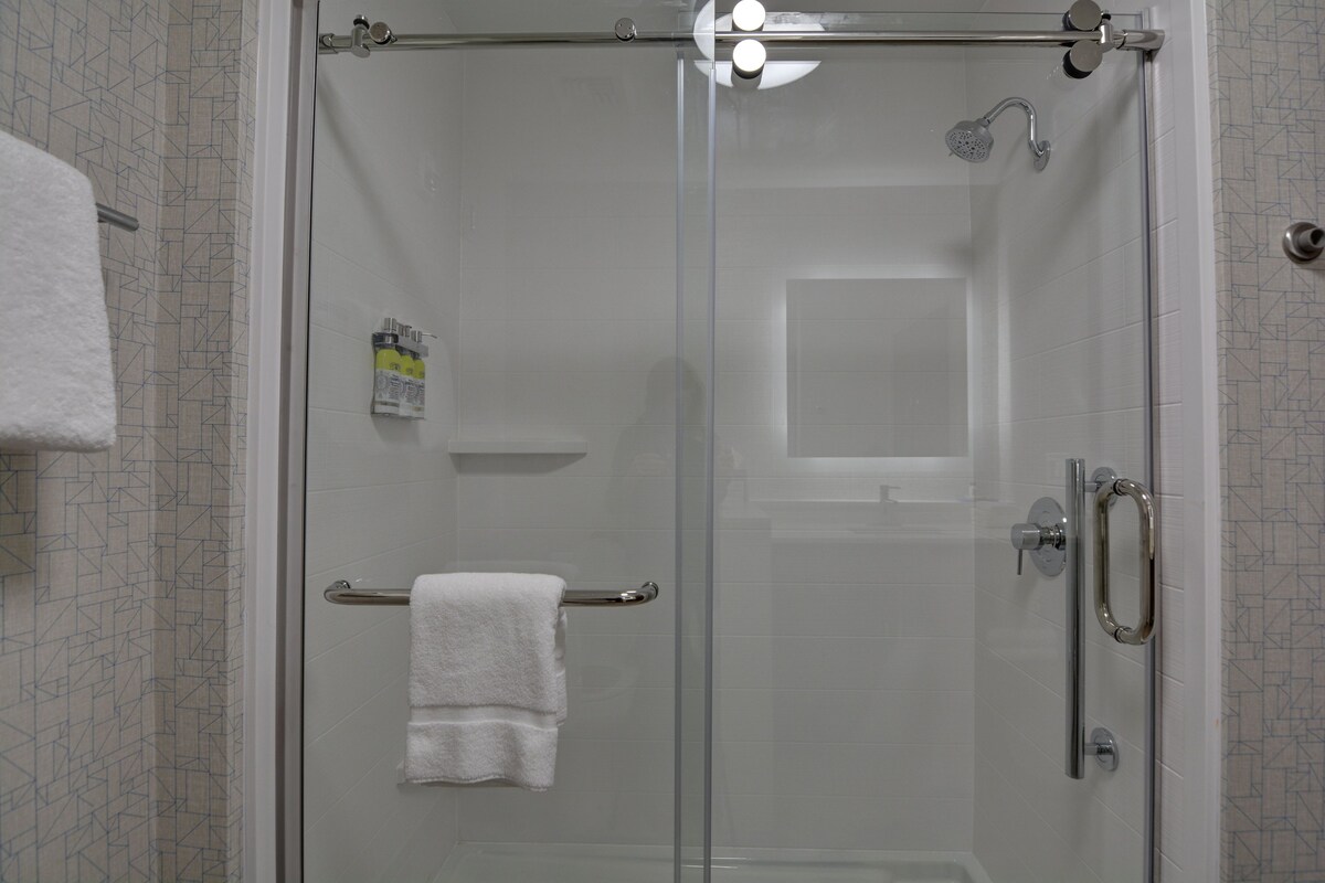 A glass-enclosed shower stall with a white towel on a towel rack, a handheld showerhead, and a bottle of shampoo.