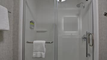 Cabina de ducha con paredes de vidrio, una toalla blanca en un perchero, un cabezal de ducha manual y una botella de champú.