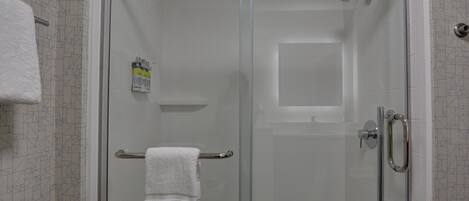 A glass-enclosed shower stall with a white towel on a towel rack, a handheld showerhead, and a bottle of shampoo.