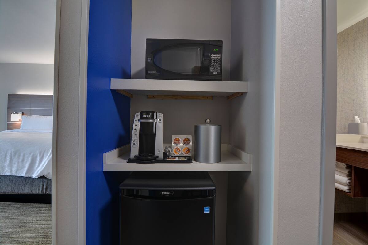Quarto de hotel compacto com cama, micro-ondas, cafeteira, mini-geladeira e prateleira pequena com itens de higiene pessoal.