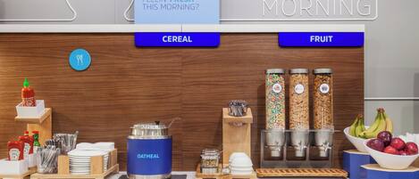 A breakfast buffet with labeled sections for cereal and fruit, including a blue container for oatmeal and jars of cereal.