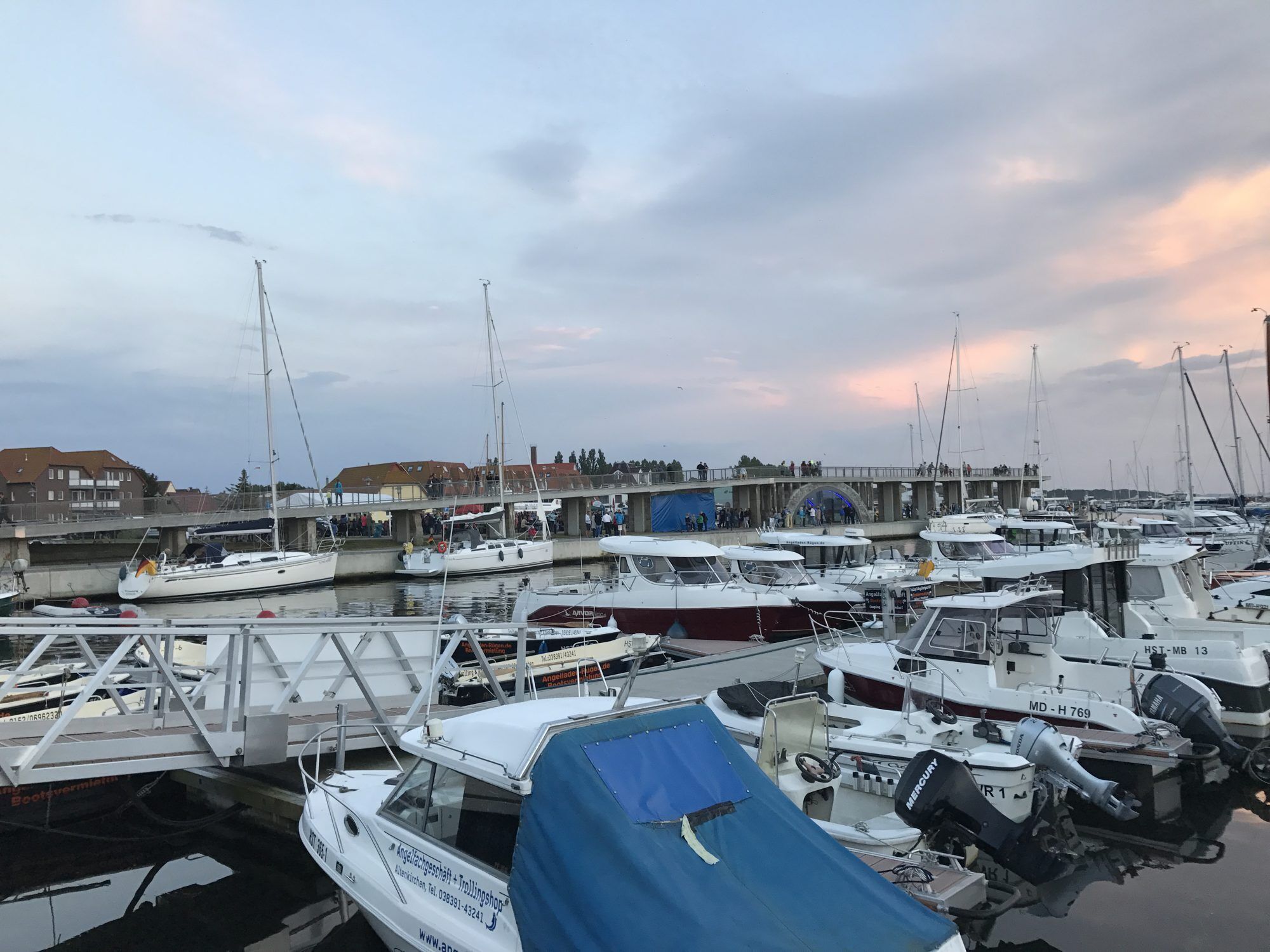 Een jachthaven met meerdere boten afgemeerd, een overdekte promenade en een brug met mensen erop.