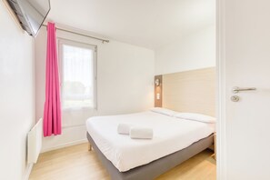A hotel room with a bed, a window with pink curtains, a bedside table, and a wall-mounted light. -  Fasthotel Roissy CDG Sud Claye-Souilly (Claye-Souilly)