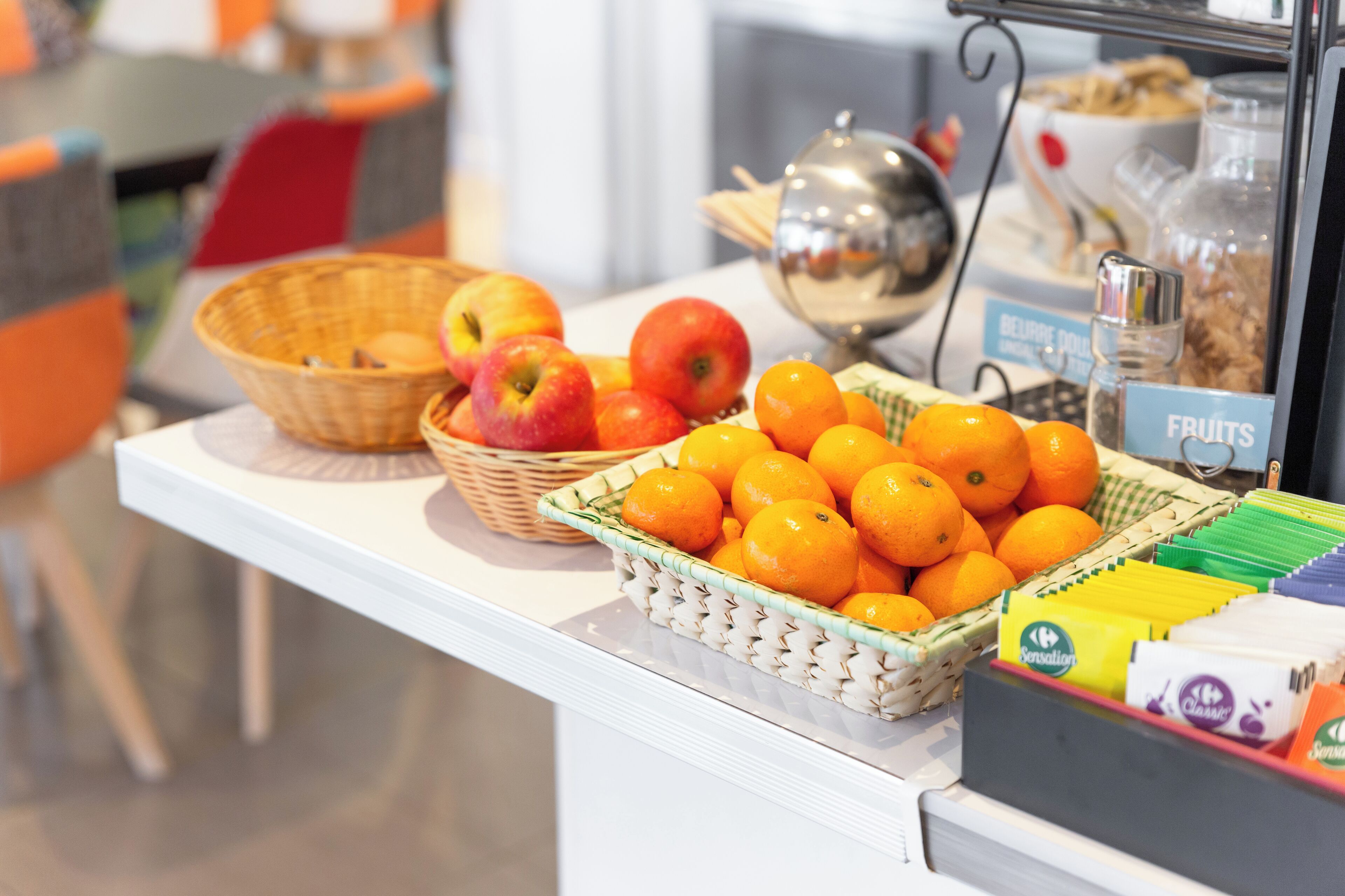 Exposição de frutas com cestos de maçãs e laranjas, uma placa com a inscrição 'FRUITS' e um recipiente com saquinhos de açúcar.