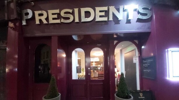 Um restaurante chamado 'PRESIDENTS' com uma placa acima da entrada, uma lousa anunciando 'Aberto para o almoço hoje!', e duas plantas em vasos na entrada.