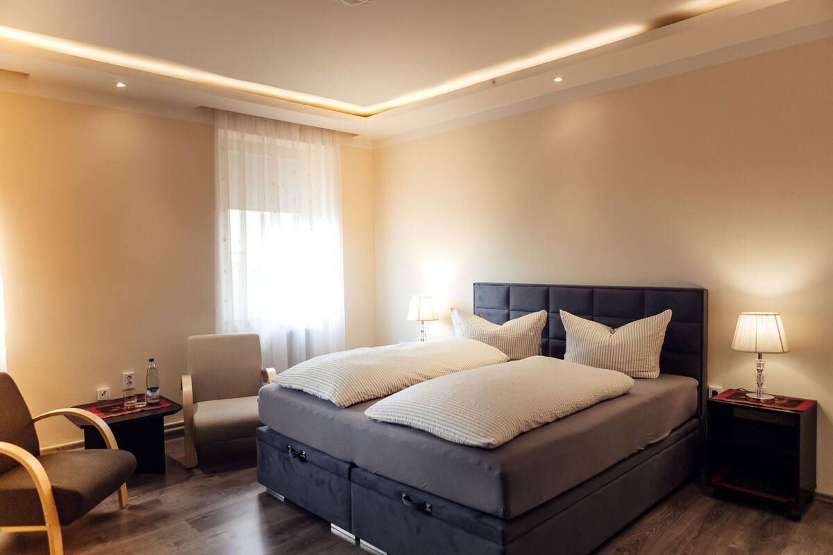 A modern hotel room with a large bed, two bedside tables with lamps, a chair, and a window with sheer curtains.