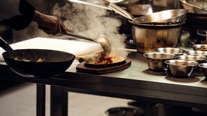 A kitchen with stainless steel cookware, a person stirring a pan, and steam rising from the cooking. - Hotel & Restaurant Peking (Riesa)