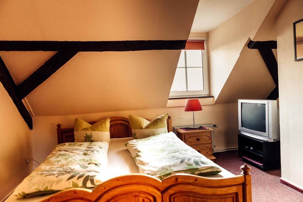 A bedroom with a sloped ceiling, a bed with floral bedding, a bedside table with a lamp, and a television.