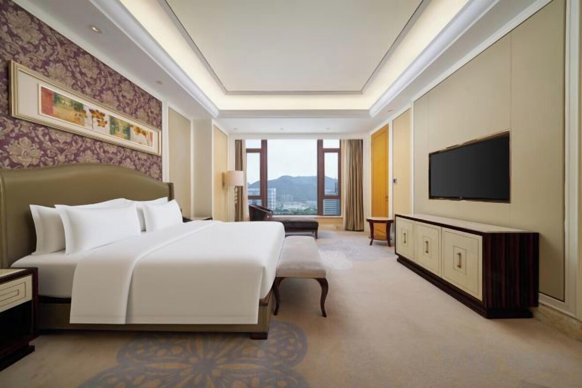Ein Hotelzimmer mit einem großen Bett, einem an der Wand befestigten Fernseher und Blick auf die Stadt.