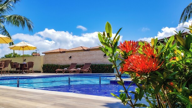 A swimming pool with lounge chairs and umbrellas, surrounded by palm trees and a building with a tiled roof.